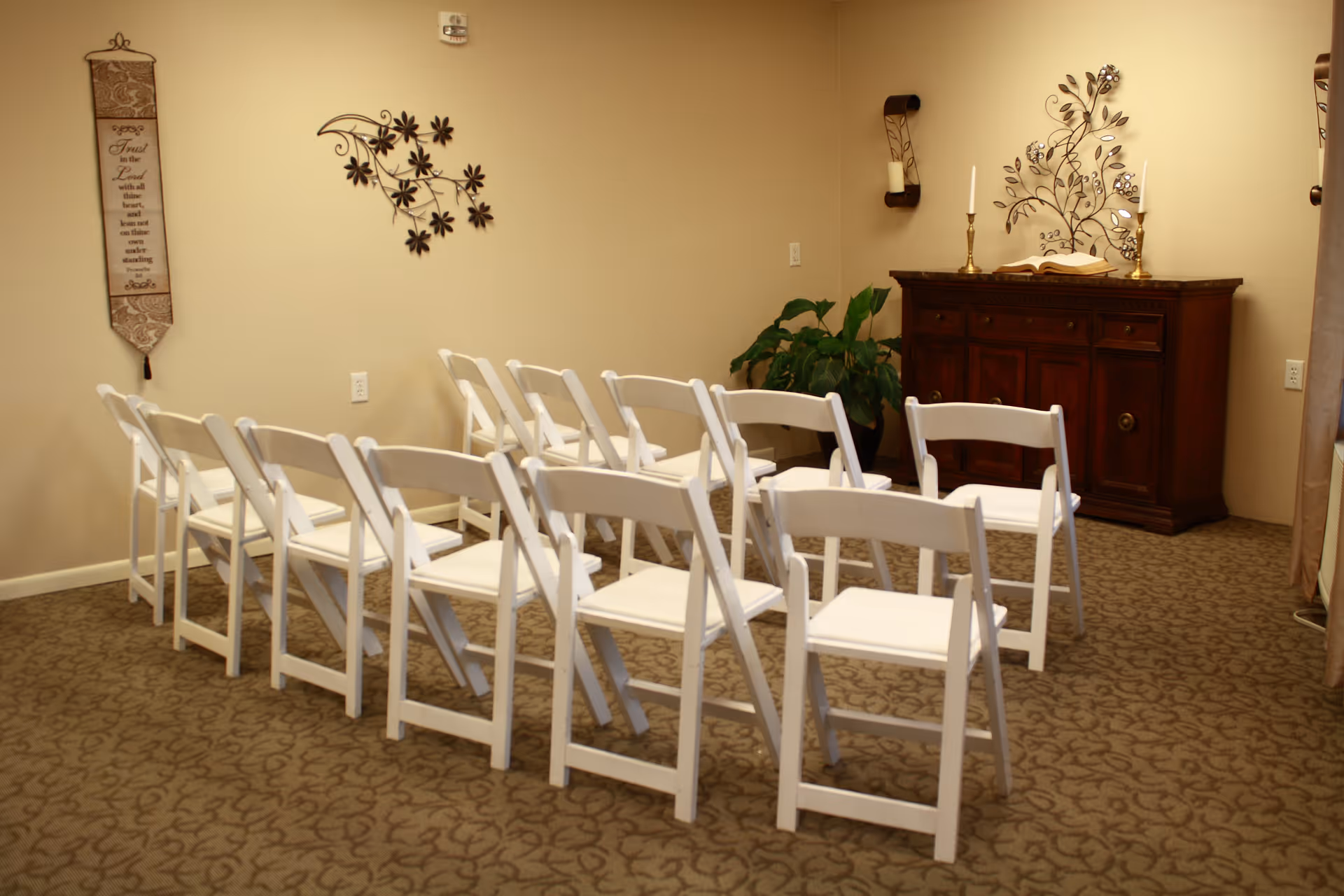 A small room with two rows of white folding chairs facing a wooden cabinet with decorative items including candles and a metal wall art piece. The room has beige walls and carpet with a floral pattern. A plant is placed near the cabinet, and a wall hanging with text is visible on the left side.