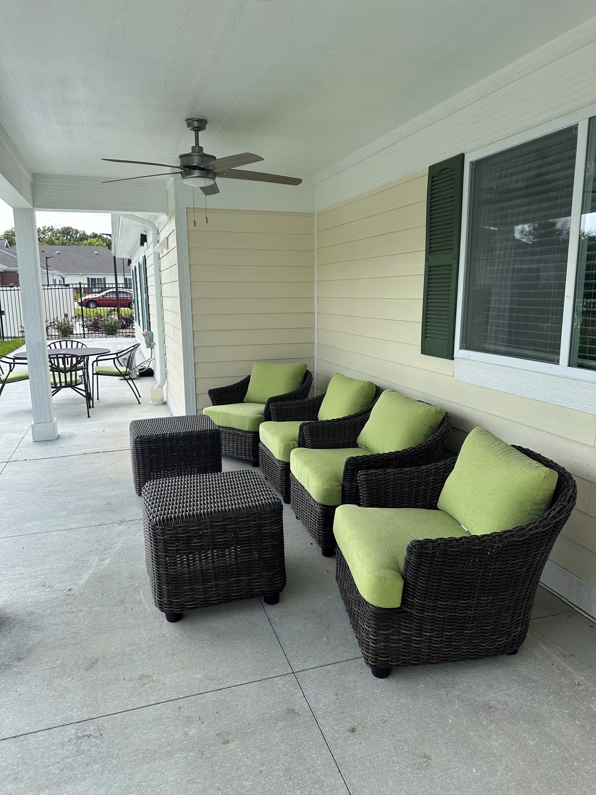 Covered outdoor patio area with four dark wicker chairs featuring light green cushions and two matching wicker ottomans. A ceiling fan is mounted on the white ceiling above. In the background, there is a round table with four chairs and a view of a fenced yard and neighboring houses.