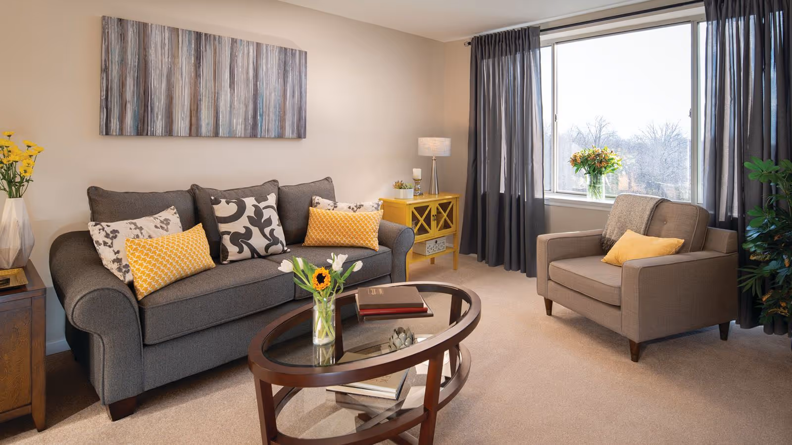 Sunlit living room with a gray sofa and matching armchair, an oval glass coffee table, yellow accent pillows and a large window with curtains.