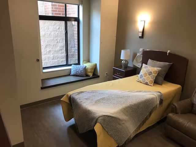 A simple single-occupancy bedroom with a single bed made with yellow bedding, a bedside table and lamp, a window seat with cushions, and a recliner.