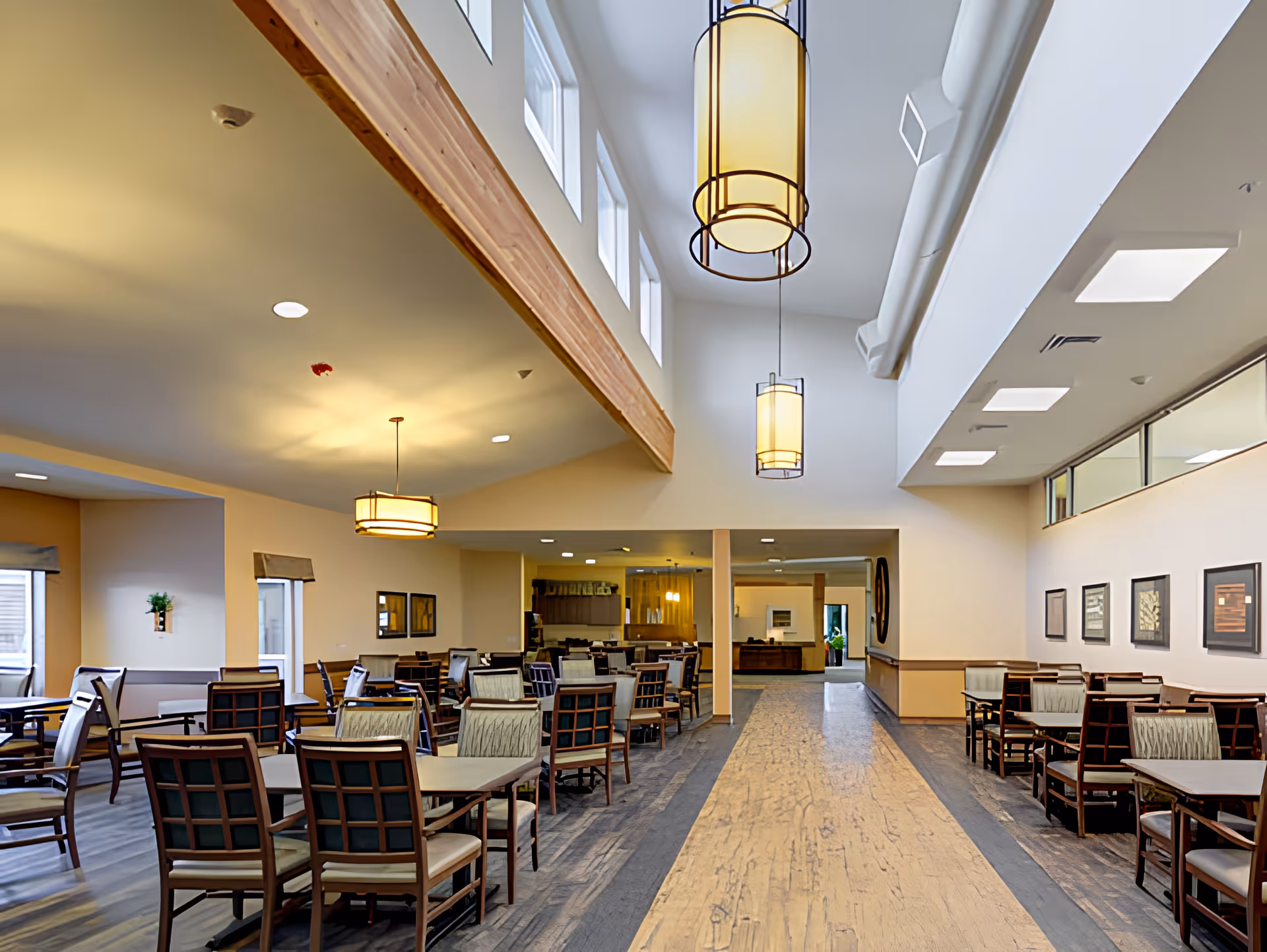 A spacious dining area in a senior living facility with multiple tables and chairs arranged neatly. The room features high ceilings with large hanging light fixtures, windows near the ceiling allowing natural light, and framed artwork on the walls. The floor has a combination of wood and a lighter central pathway.