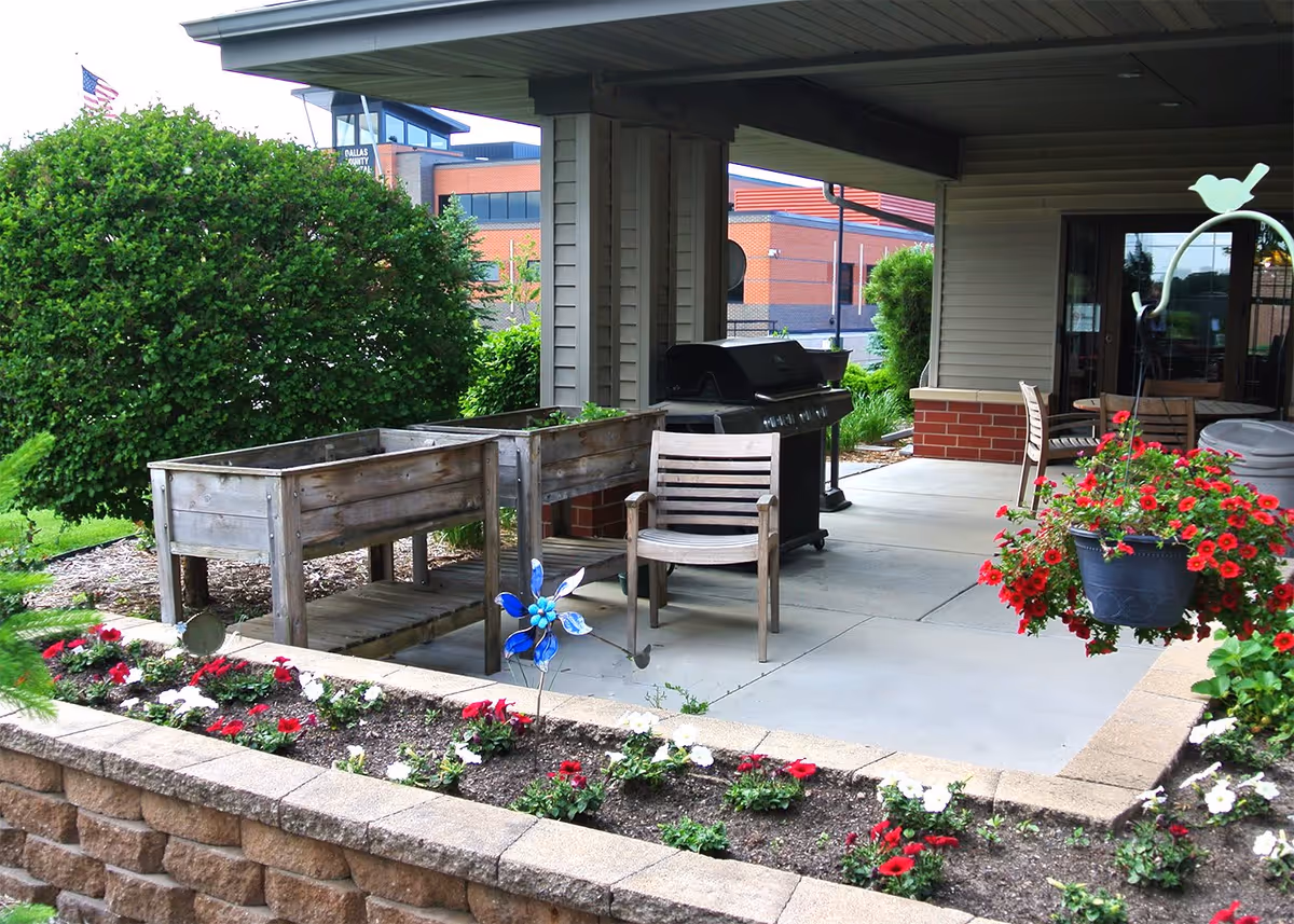 Covered outdoor patio area with wooden chairs, a barbecue grill, raised garden beds, and a flower bed with red and white flowers in front. There is a hanging flower pot with red flowers on the right side and a large green bush in the background.