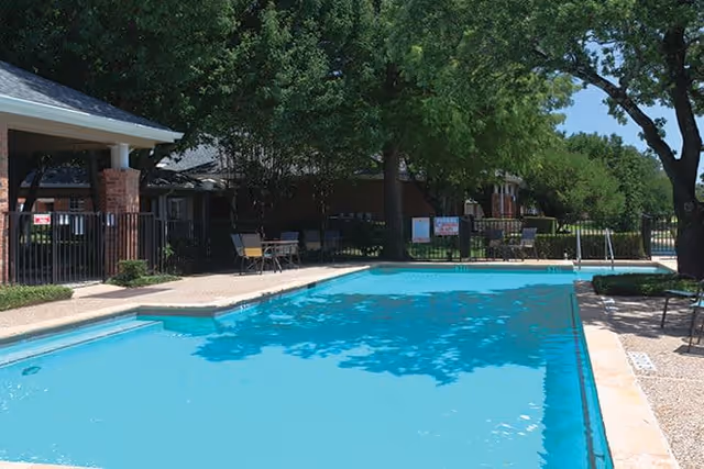 Outdoor swimming pool surrounded by trees, chairs, and a fence with a covered seating area on the left side under a roof.