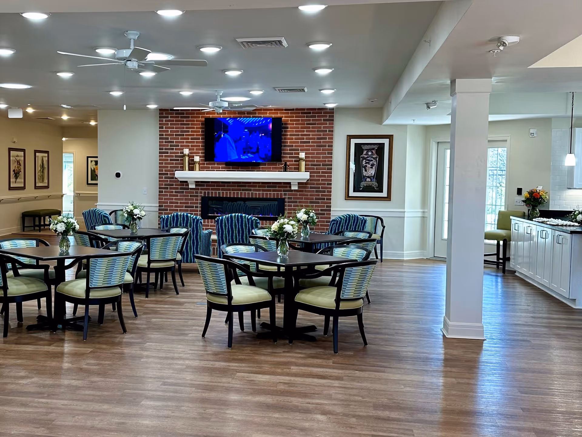 A spacious dining room with several round tables and chairs arranged neatly. Each table has a vase with white flowers. The room features a brick fireplace with a mounted flat-screen TV above it. The walls are decorated with framed artwork, and the floor is wooden. Ceiling fans and recessed lighting are visible on the ceiling. To the right, there is a kitchen area with white cabinets and a countertop with flowers on it.