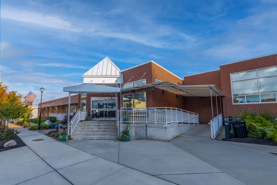 Front entrance of a brick senior living building with concrete steps and a wheelchair ramp under a canopy, landscaped walkways and an American flag.