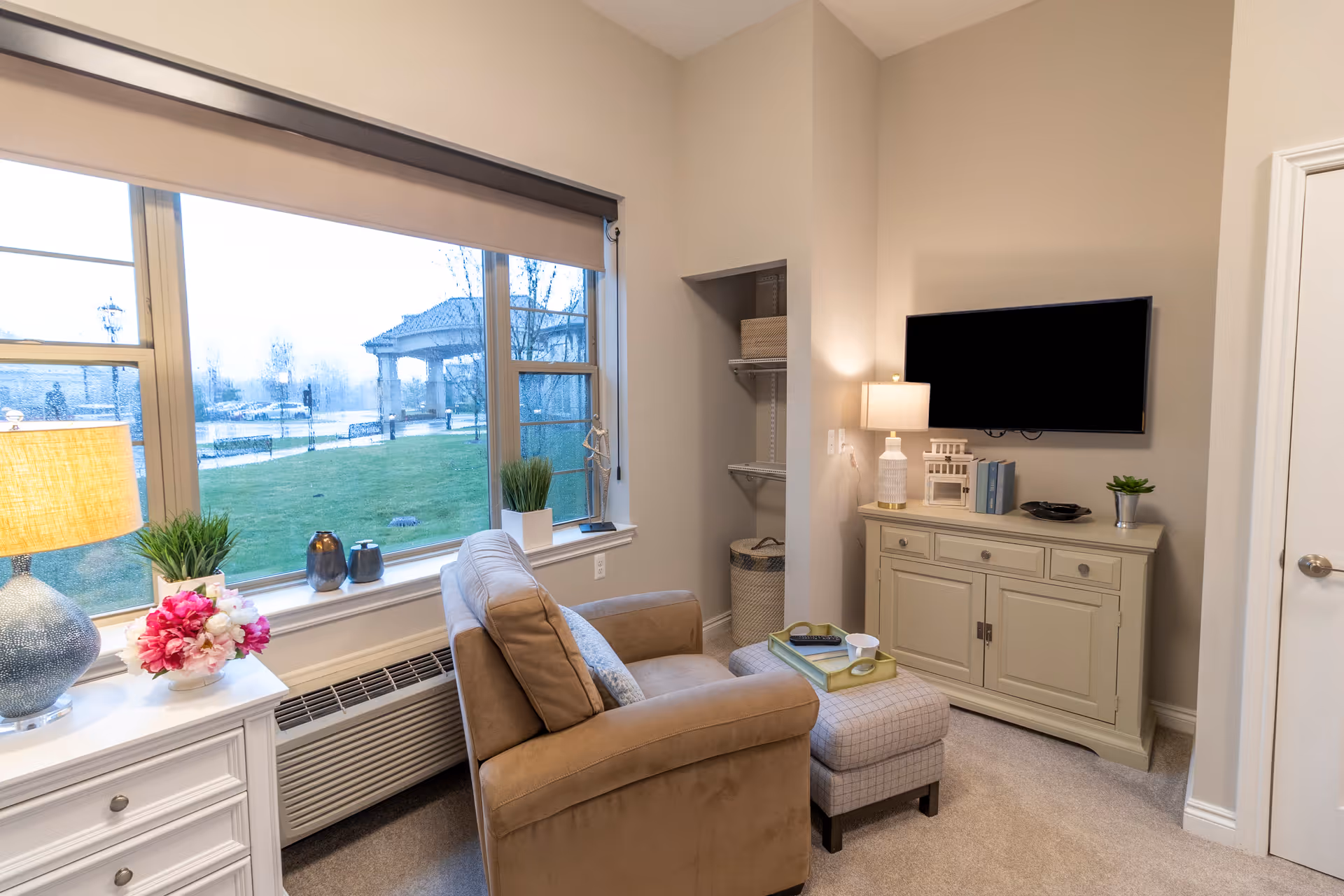 Cozy living area with an armchair and ottoman facing a wall-mounted TV, a cabinet, large window overlooking a lawn, and lamps and decorative plants.