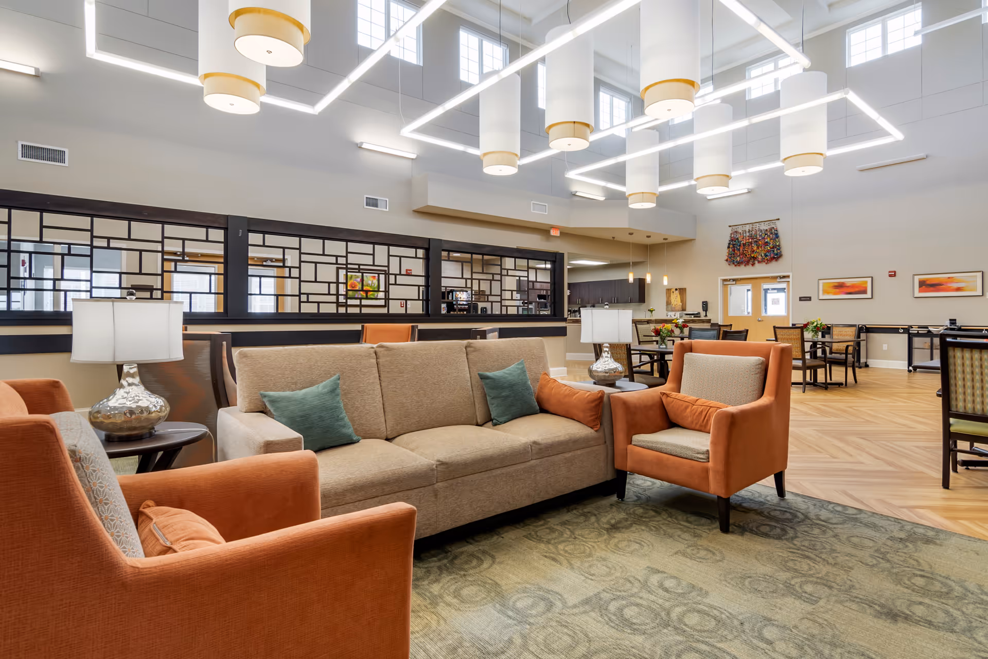 A spacious and well-lit common area with a beige sofa and two orange armchairs arranged around a small table with a lamp. The room features high ceilings with large modern light fixtures, a patterned carpet, and a wooden floor. In the background, there are dining tables and chairs, decorative wall art, and a partition with a geometric design separating the seating area from another room.
