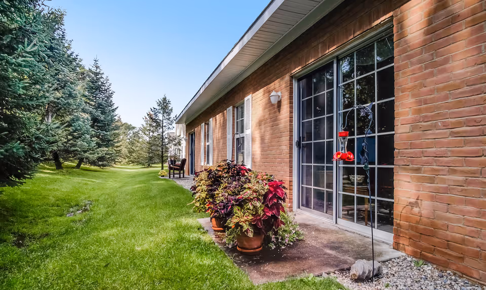 Outdoor view of a brick building with sliding glass doors and windows, surrounded by green grass and trees. There are potted plants near the building and a bird feeder hanging in front of the glass door.