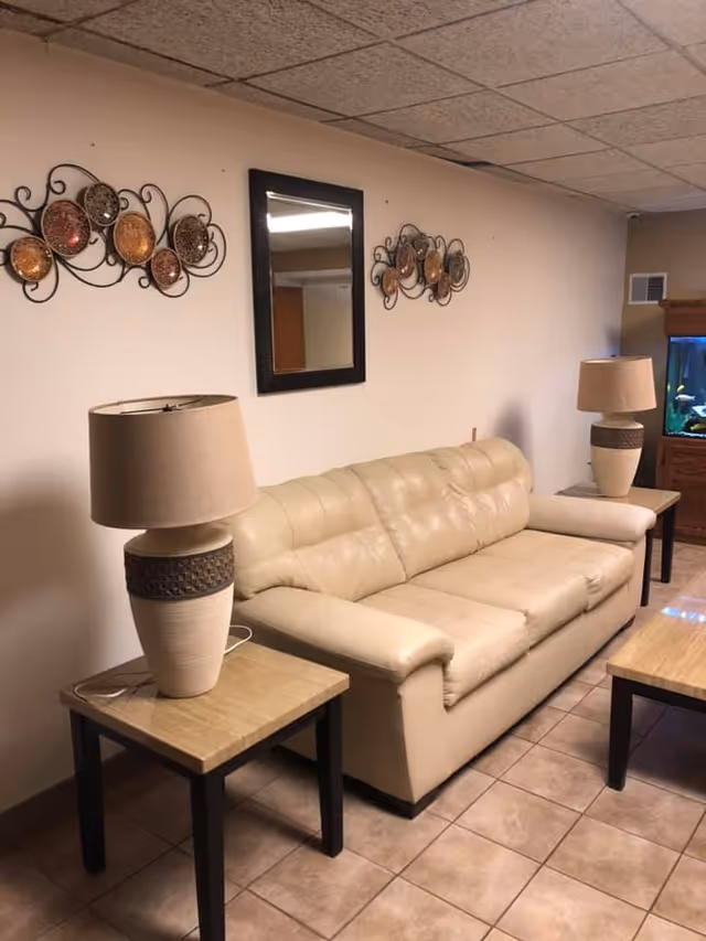 A beige leather sofa with two matching side tables, each holding a beige lamp with a textured band. Above the sofa is a rectangular mirror flanked by decorative metal wall art with circular designs. To the right, there is a wooden cabinet with a fish tank on top. The floor is tiled and the walls are light-colored.