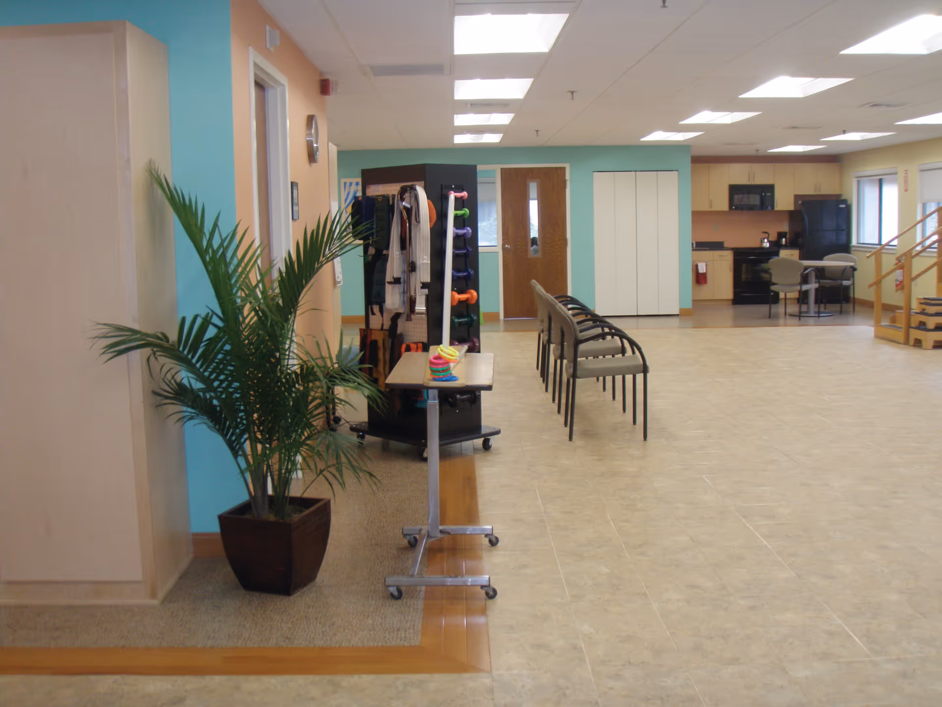 Interior view of a senior living facility room with a row of chairs, a small table with colorful rings, a plant in a pot, exercise equipment including dumbbells and resistance bands, and a kitchenette area with a microwave, refrigerator, and table with chairs in the background.
