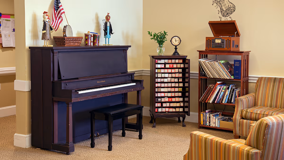 A cozy corner of a living room featuring a dark upright piano with a bench, two decorative rooster figurines, and an American flag on top. Next to the piano is a small cabinet with many labeled drawers and a clock on top. To the right, there is a wooden shelf holding vinyl records, books, and a vintage record player. Two striped upholstered armchairs are partially visible in the foreground.