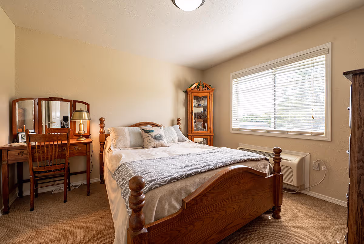 A cozy bedroom with a wooden bed frame, a neatly made bed with white and gray bedding, a wooden vanity with a mirror and chair, a lamp, and a wooden corner cabinet. There is a large window with blinds letting in natural light, beige walls, and carpeted floor.