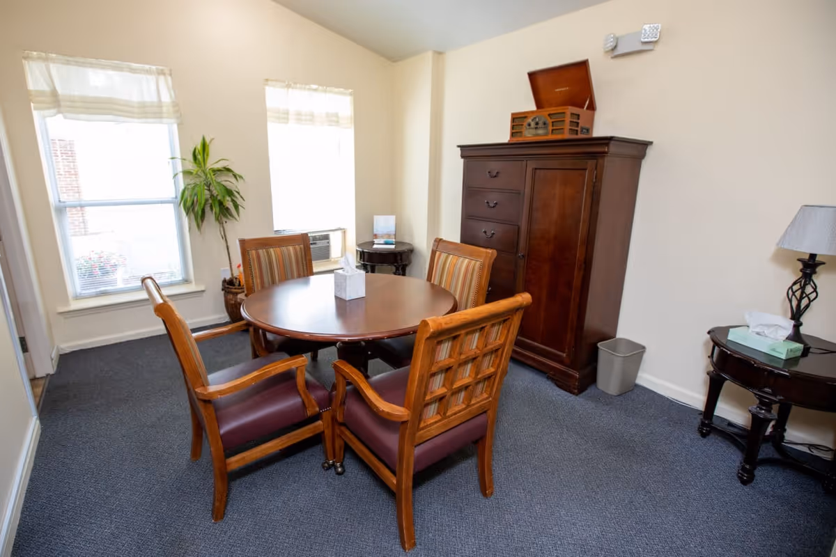 Small interior dining area with a round wooden table surrounded by four chairs, a tall wooden cabinet, windows, and a side table with a lamp.