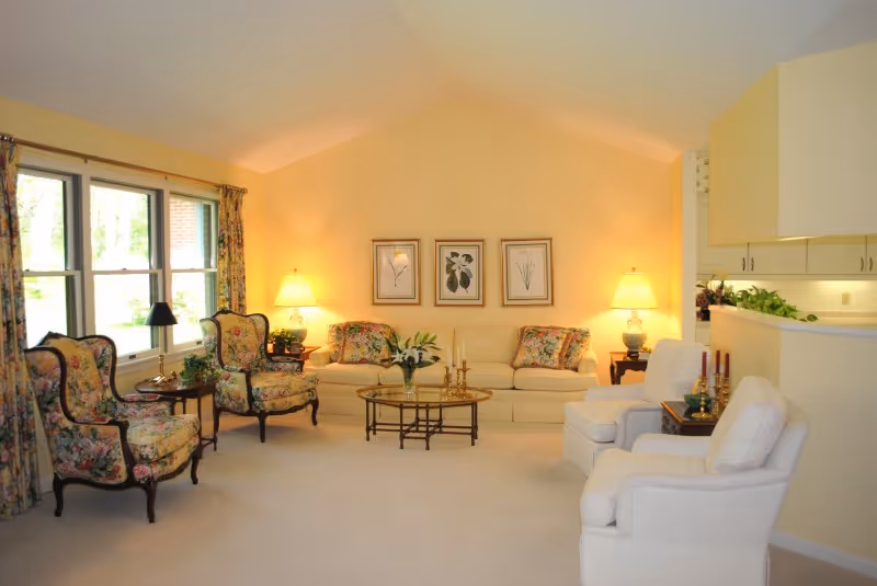 A cozy living room with cream-colored walls and carpet, featuring a cream sofa with floral cushions, two floral upholstered armchairs, two white armchairs, a glass coffee table with decorative items, two side tables with lamps, and three botanical prints on the wall. Large windows with floral curtains allow natural light into the room.