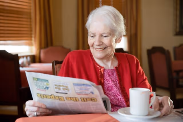 An elderly woman with white hair wearing a red cardigan and pink patterned shirt is sitting at a table in a dining area, smiling while reading a newspaper. A white cup and saucer are on the table in front of her, and there are wooden chairs and a window with blinds in the background.