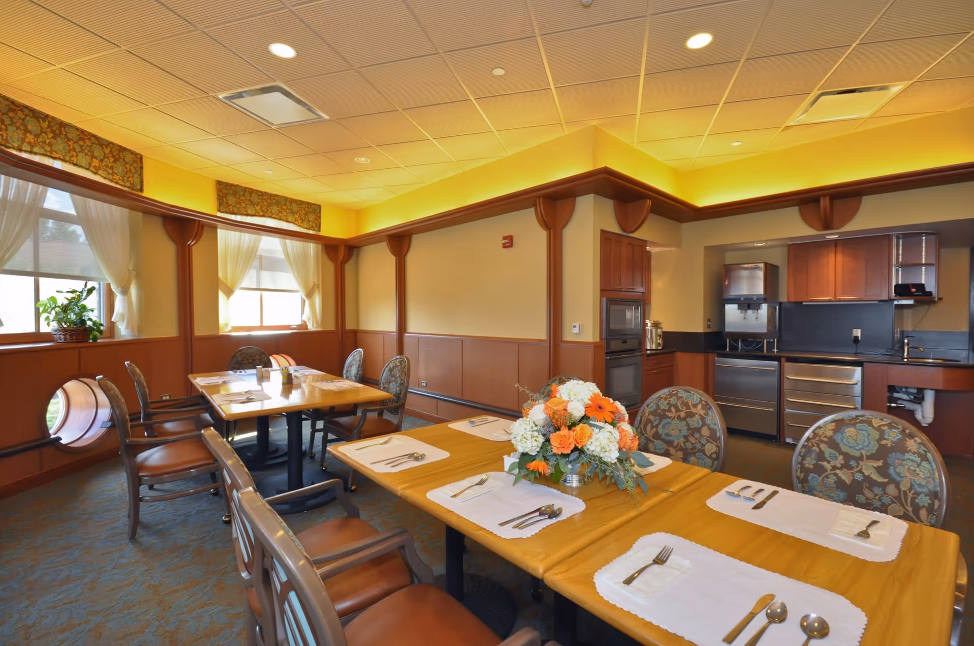 A dining room in St. Joseph Manor with wooden tables set with placemats, silverware, and a floral centerpiece. The room features patterned chairs, large windows with sheer curtains, warm yellow lighting, and a small kitchen area with cabinets and appliances in the background.
