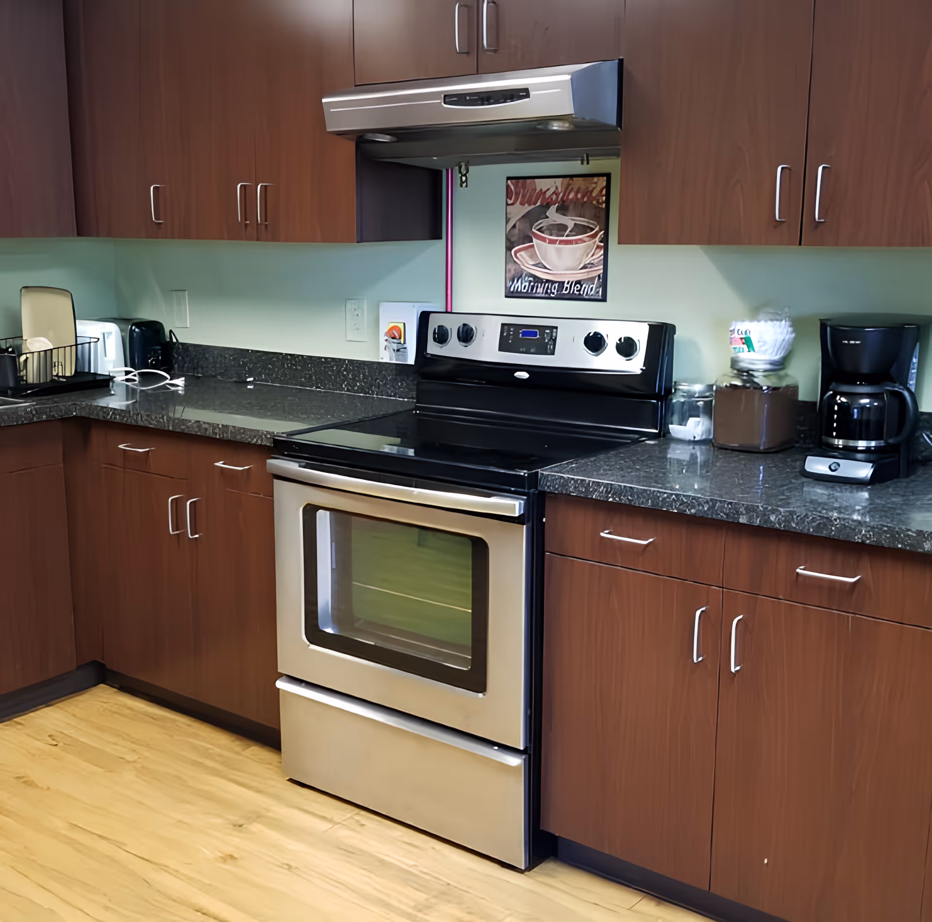 A kitchen area with wooden cabinets, a stainless steel electric stove with oven, a coffee maker, and various kitchen items on the dark granite countertop. There is a framed picture of a steaming coffee cup on the wall above the stove.
