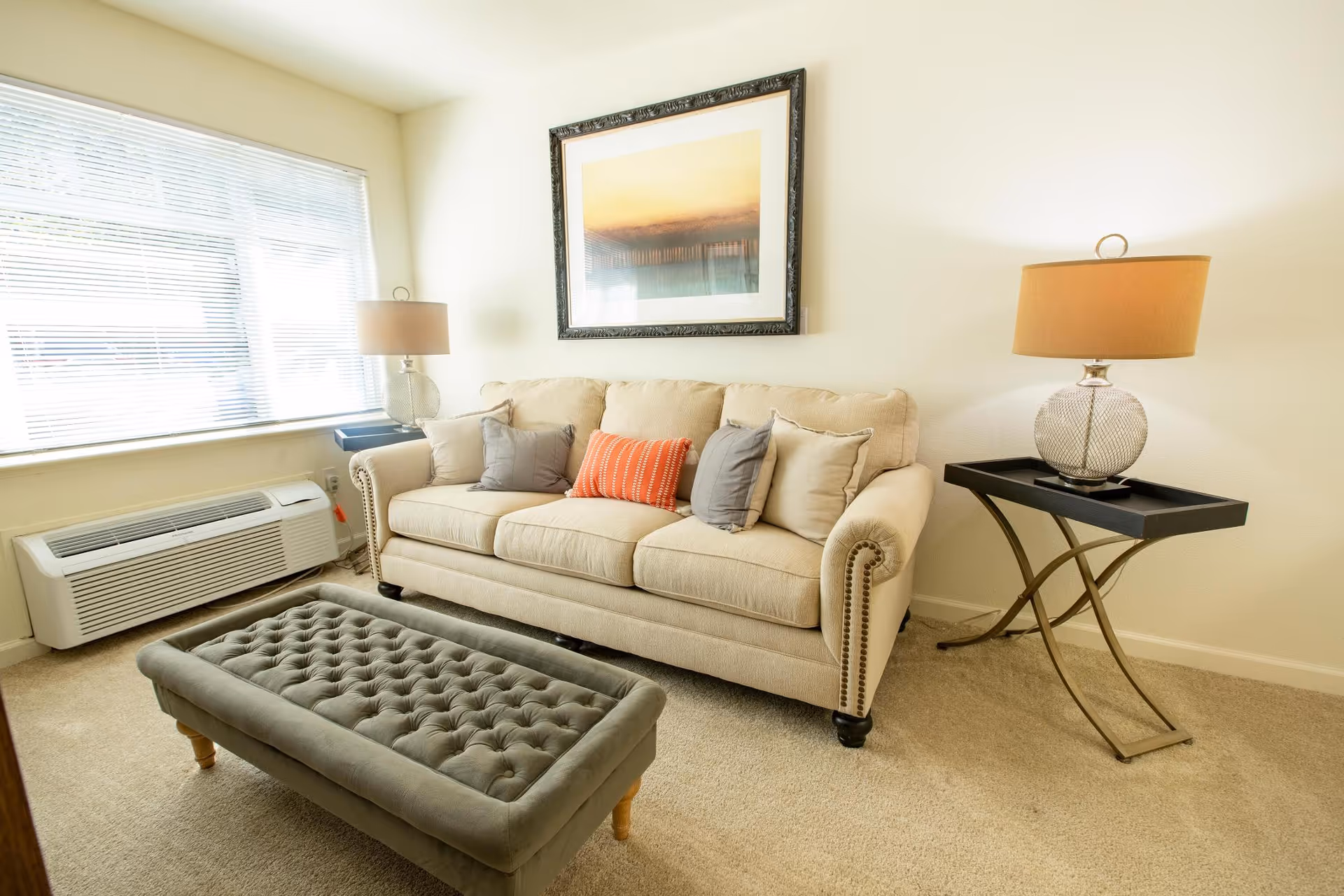 A bright living room with a beige sofa adorned with five cushions in gray, beige, and orange colors. In front of the sofa is a tufted gray ottoman. On either side of the sofa are side tables with glass lamps. A large window with blinds allows natural light to fill the room. A framed artwork hangs on the wall above the sofa.