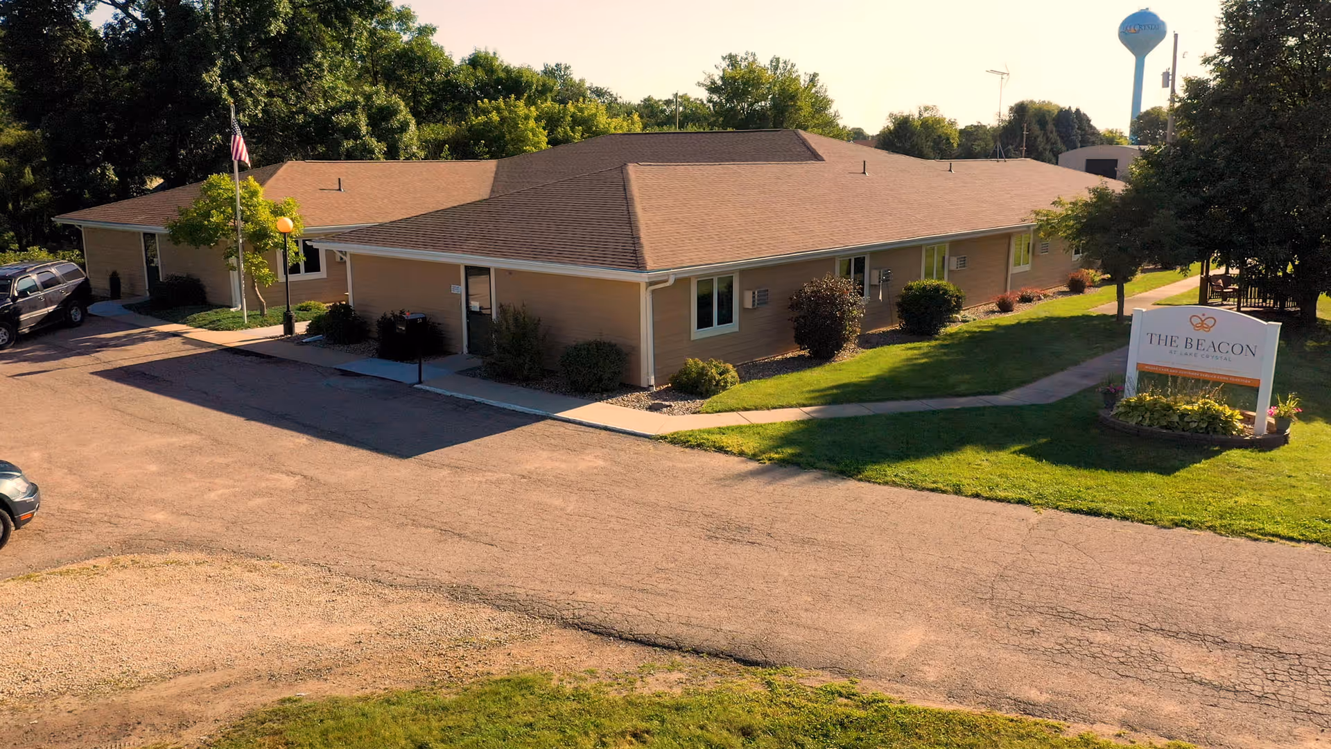 Exterior view of a single-story senior living facility building with a brown roof and beige walls, surrounded by green grass, bushes, and trees. There is a paved driveway and parking area with a few parked cars. A sign near the building reads 'The Beacon at Lake Crystal.' A water tower is visible in the background.
