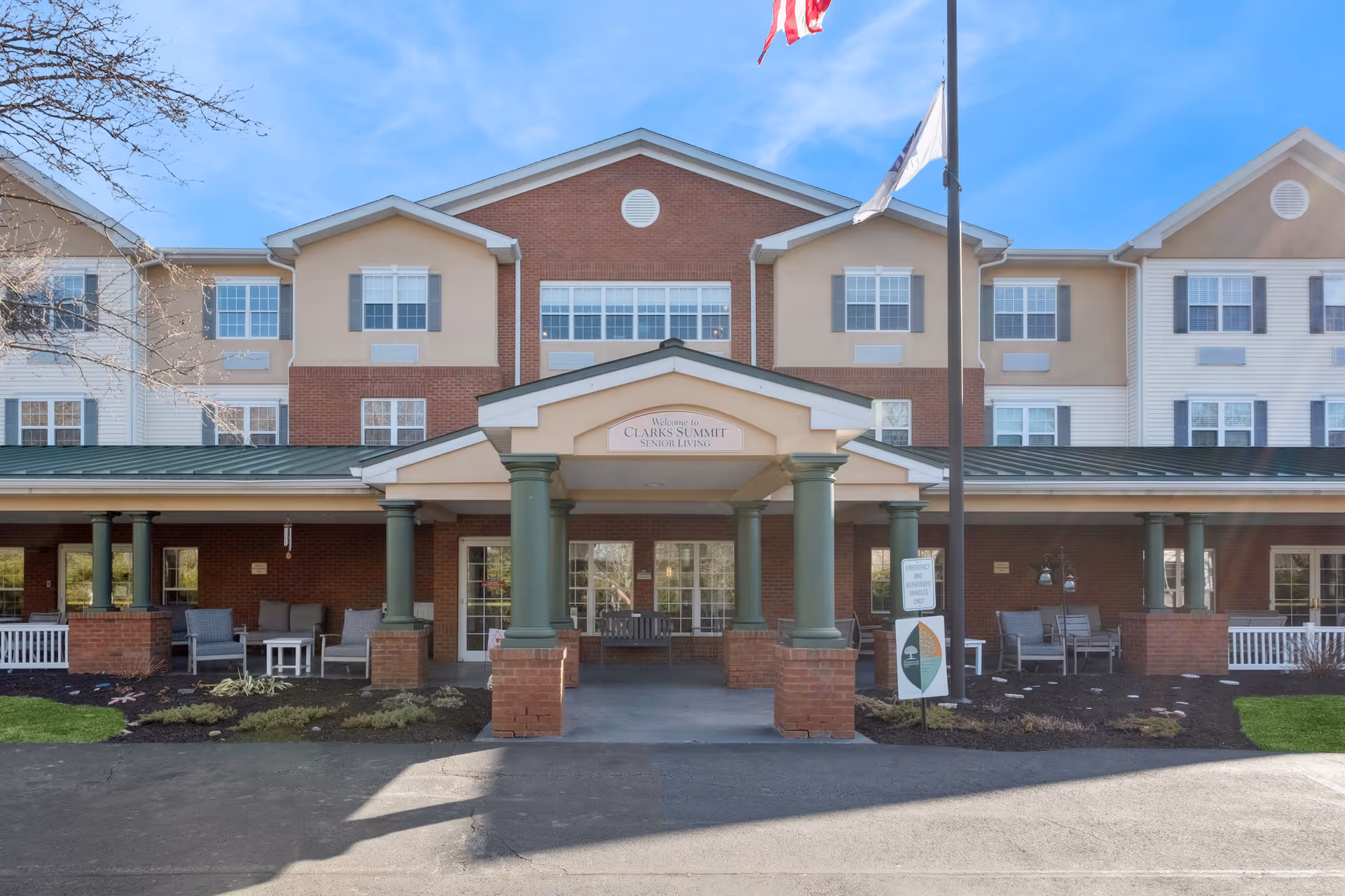 Front entrance of Clarks Summit Senior Living showing a covered portico with green columns, outdoor seating and an American flag.