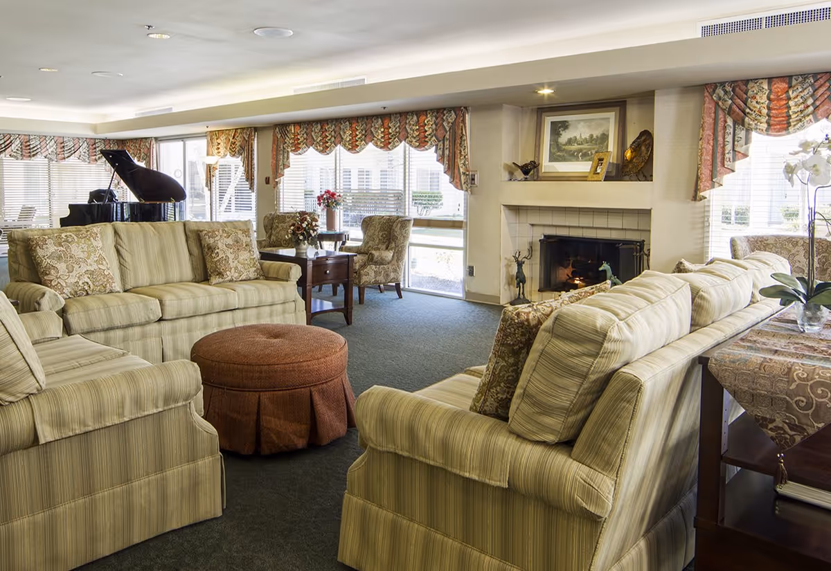 A cozy living room with two striped sofas facing each other, a round brown ottoman in the center, a fireplace with a framed picture above it, floral curtains on large windows, a grand piano in the corner, and a small table with floral arrangements and chairs near the windows.