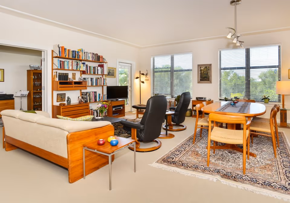 Bright open living and dining area with wooden furniture, bookshelves, leather recliners, and a dining table on a patterned rug.