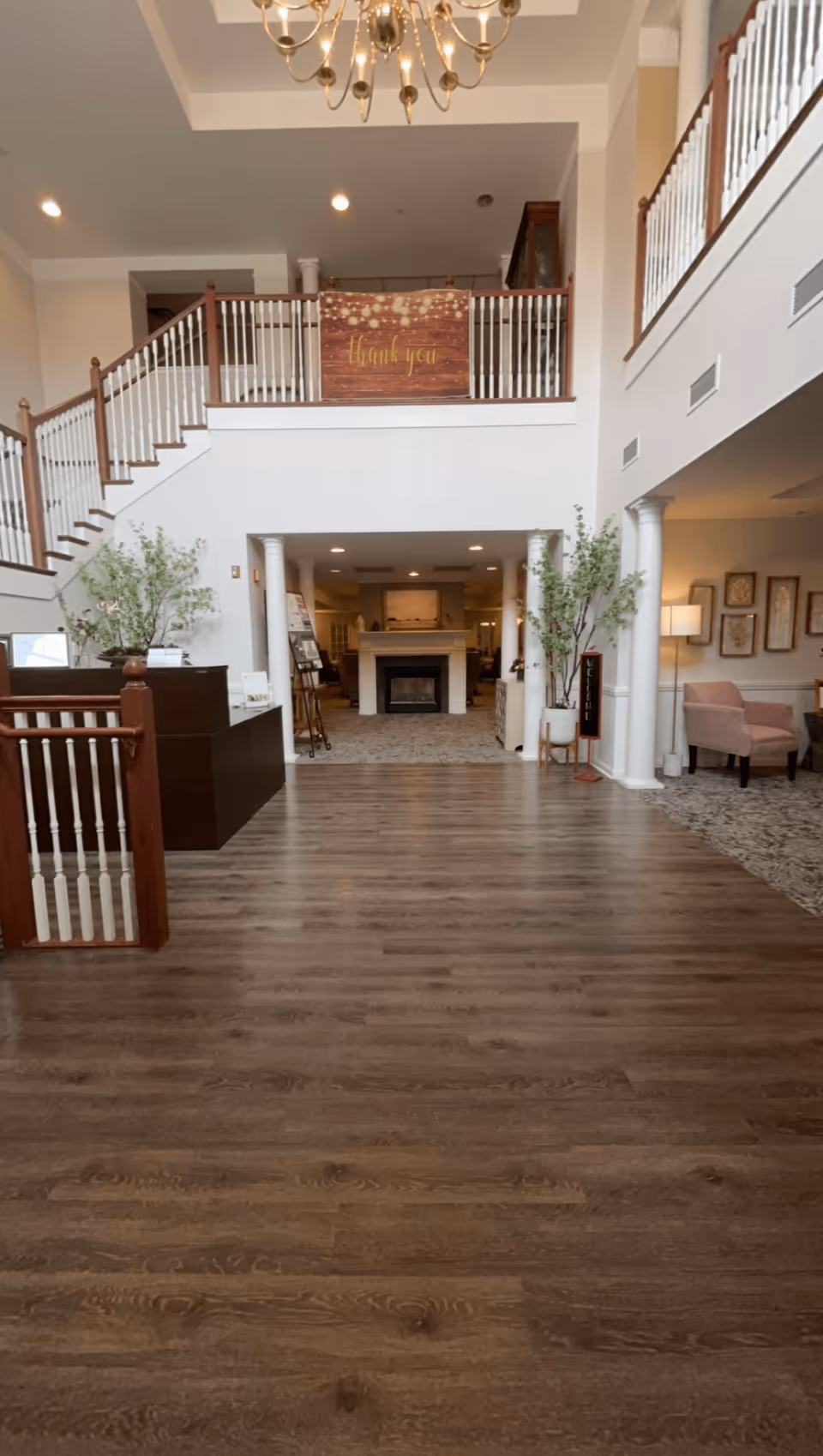 Spacious interior lobby area of a senior living facility with wooden flooring, white walls, and a high ceiling. There is a staircase with white railings and wooden banisters on the left side, and a balcony above with a 'thank you' sign. Two tall potted plants flank the entrance to a room with a fireplace. To the right, there is a seating area with chairs and framed artwork on the wall.