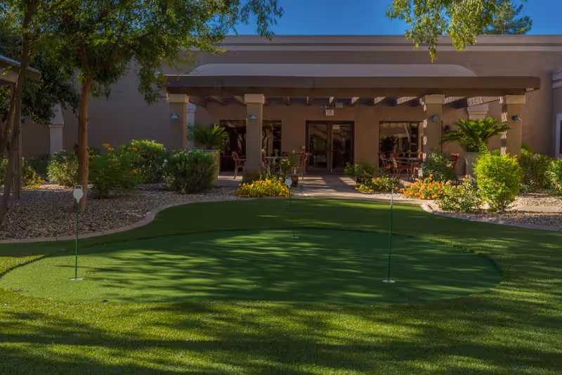 Outdoor putting green with three golf holes in front of a shaded patio area with tables and chairs, surrounded by plants and trees at Springdale Village Assisted Living Facility & Independent Living Apartments in Mesa Arizona.