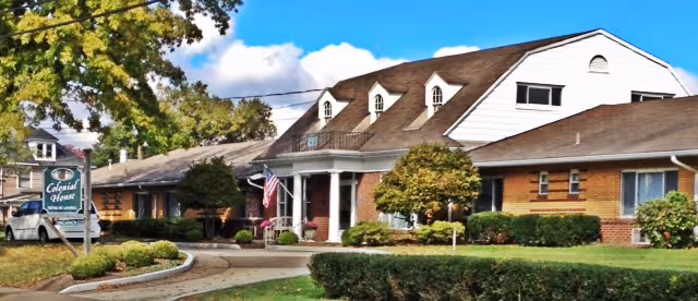 Exterior view of Colonial House Senior Independent Living facility showing a large brick building with a sloped roof, multiple windows, and a front entrance with white columns. The surrounding area includes well-maintained bushes, trees, a curved driveway, and a sign displaying the facility's name. The sky is blue with some clouds.