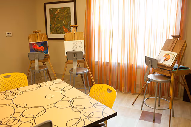 A bright room with a table featuring a white surface with black circular patterns, surrounded by yellow chairs. Three wooden easels with stools are positioned near a window with sheer orange curtains, each holding a canvas with artwork in progress. A framed botanical print hangs on the beige wall.