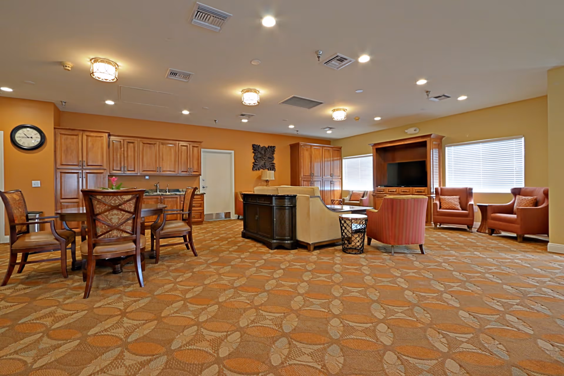 A spacious senior living common area with patterned carpet flooring, a round wooden dining table with chairs, a kitchenette with wooden cabinets, and a seating area with armchairs and a sofa facing a large wooden entertainment center with a TV. The room is well-lit with ceiling lights and has windows with blinds.
