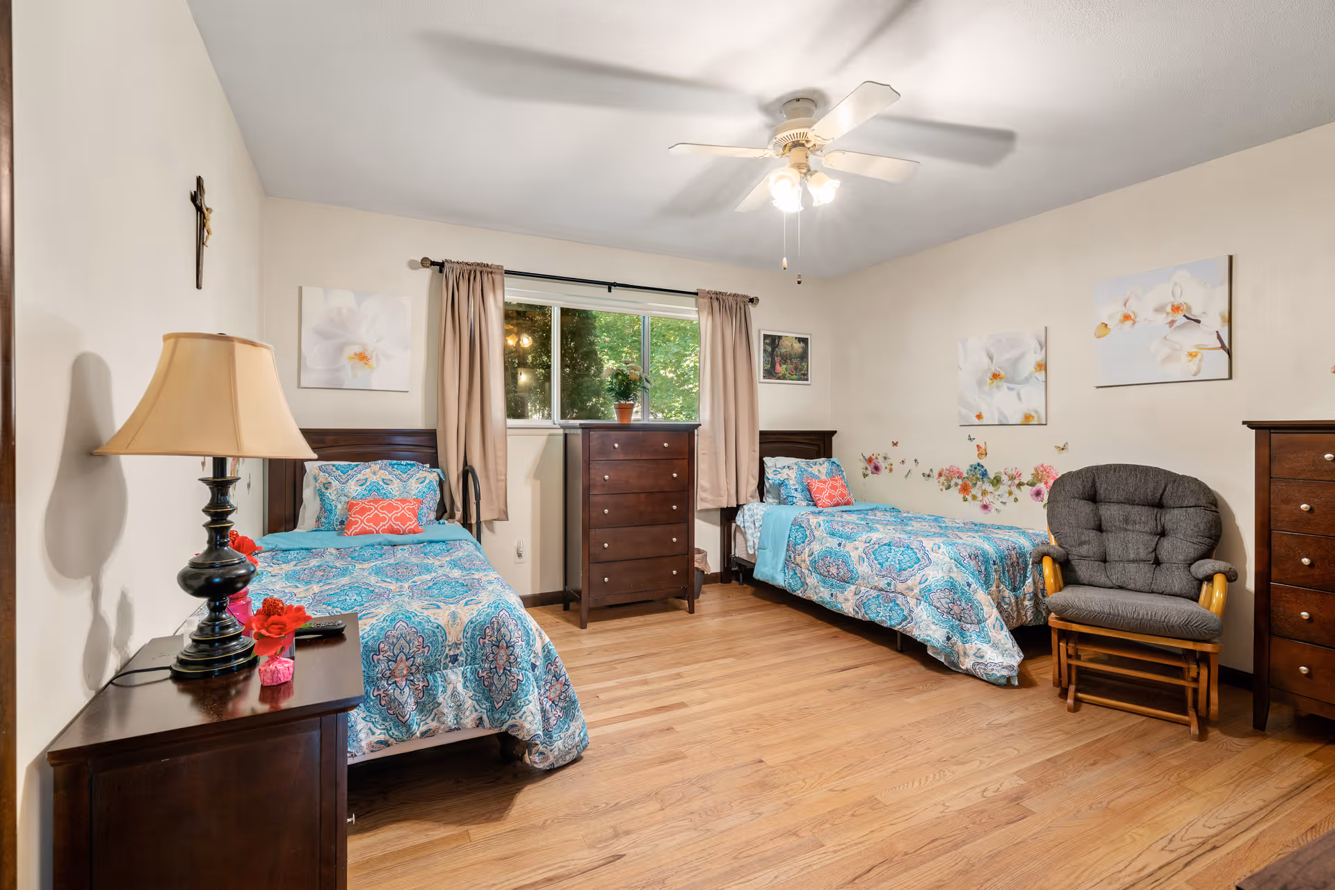 A bedroom with two twin beds featuring blue patterned bedspreads and coral accent pillows. Between the beds is a window with beige curtains and a dark wooden dresser beneath it. On the left side, there is a nightstand with a lamp and a small flower decoration. On the right side, there is a cushioned rocking chair and another dark wooden dresser. The walls are decorated with floral paintings and butterfly decals. The room has wooden flooring and a ceiling fan with lights.