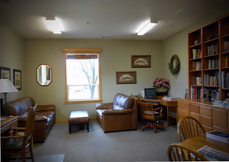 A cozy living room with two brown leather sofas, a matching ottoman, a wooden desk with a computer and chair, a large wooden bookshelf filled with books, a round wooden table with chairs, and a window with a view of a tree outside. The walls are decorated with framed pictures and a wreath.