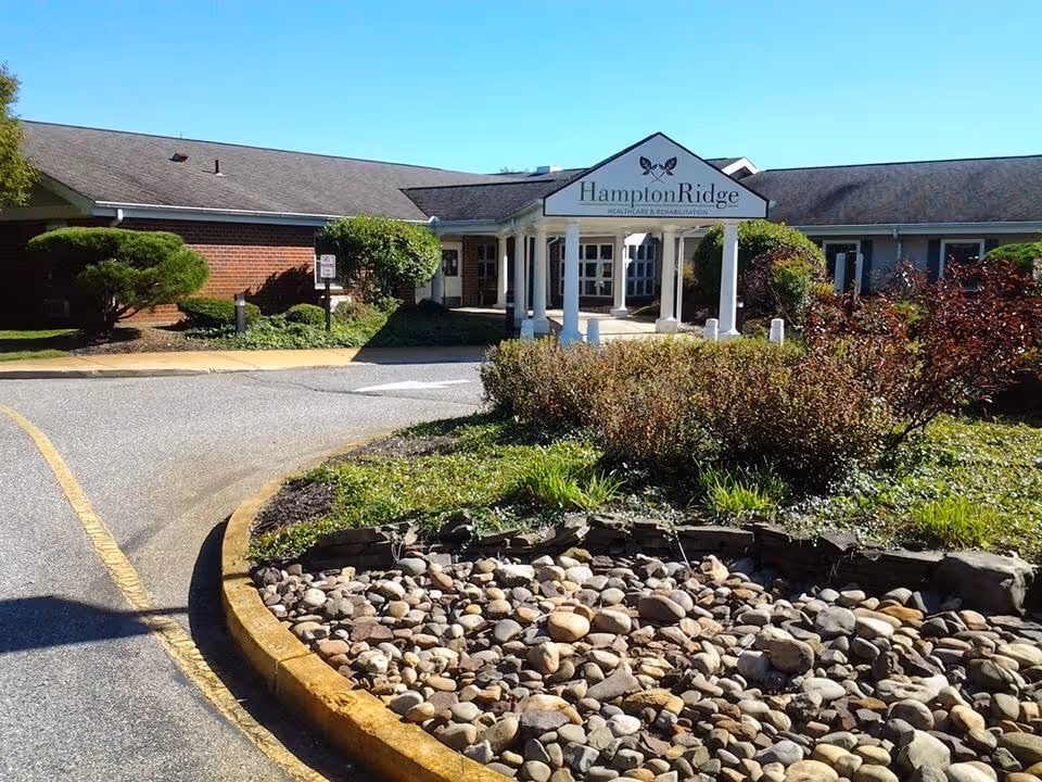 Exterior view of Hampton Ridge Healthcare & Rehabilitation building with a driveway, landscaped garden featuring rocks and bushes, and a clear blue sky.