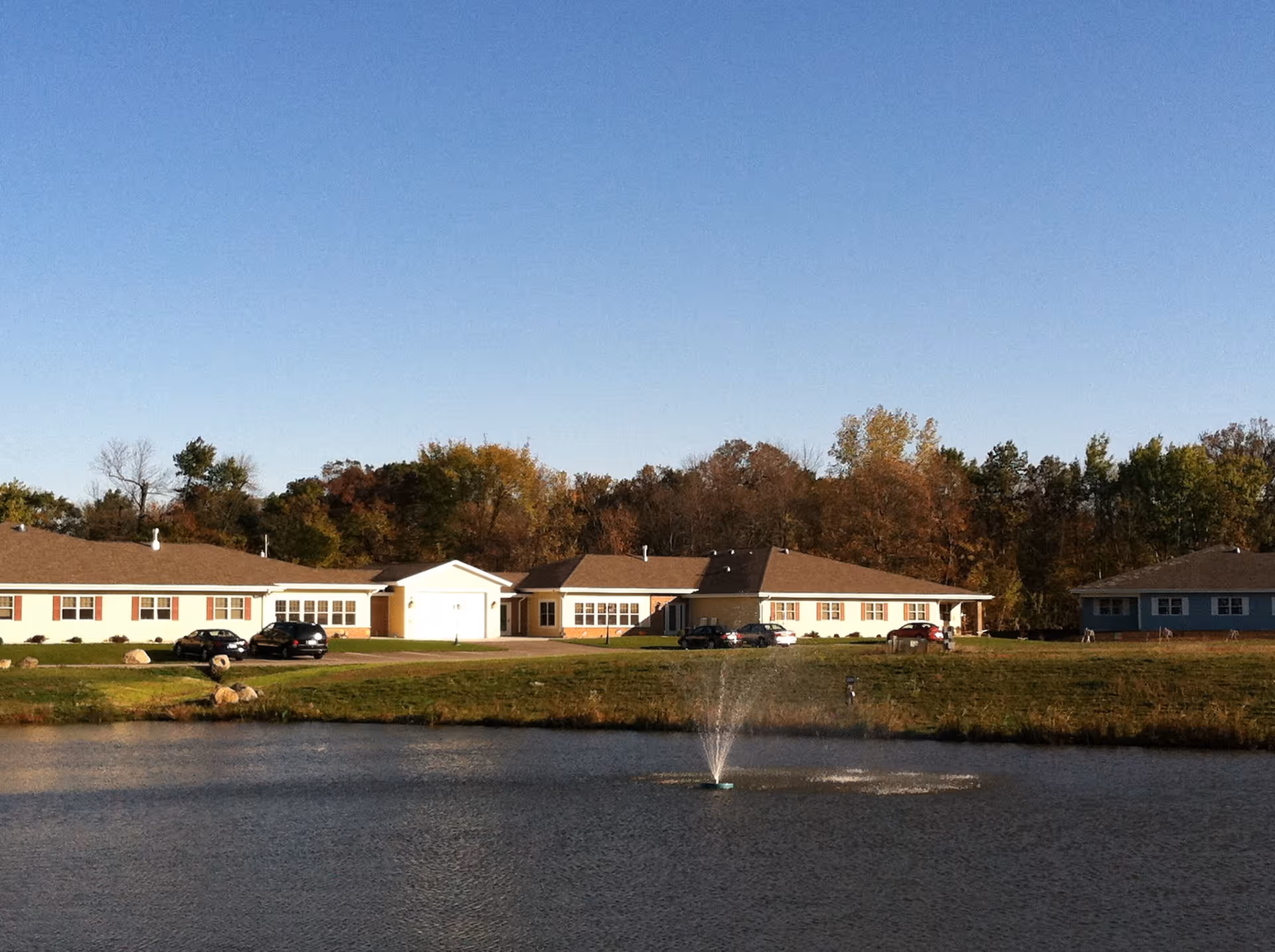 Exterior view of Maplewood Cottage senior living facility with multiple single-story buildings, a pond with a water fountain in the foreground, and trees with autumn foliage in the background under a clear blue sky.