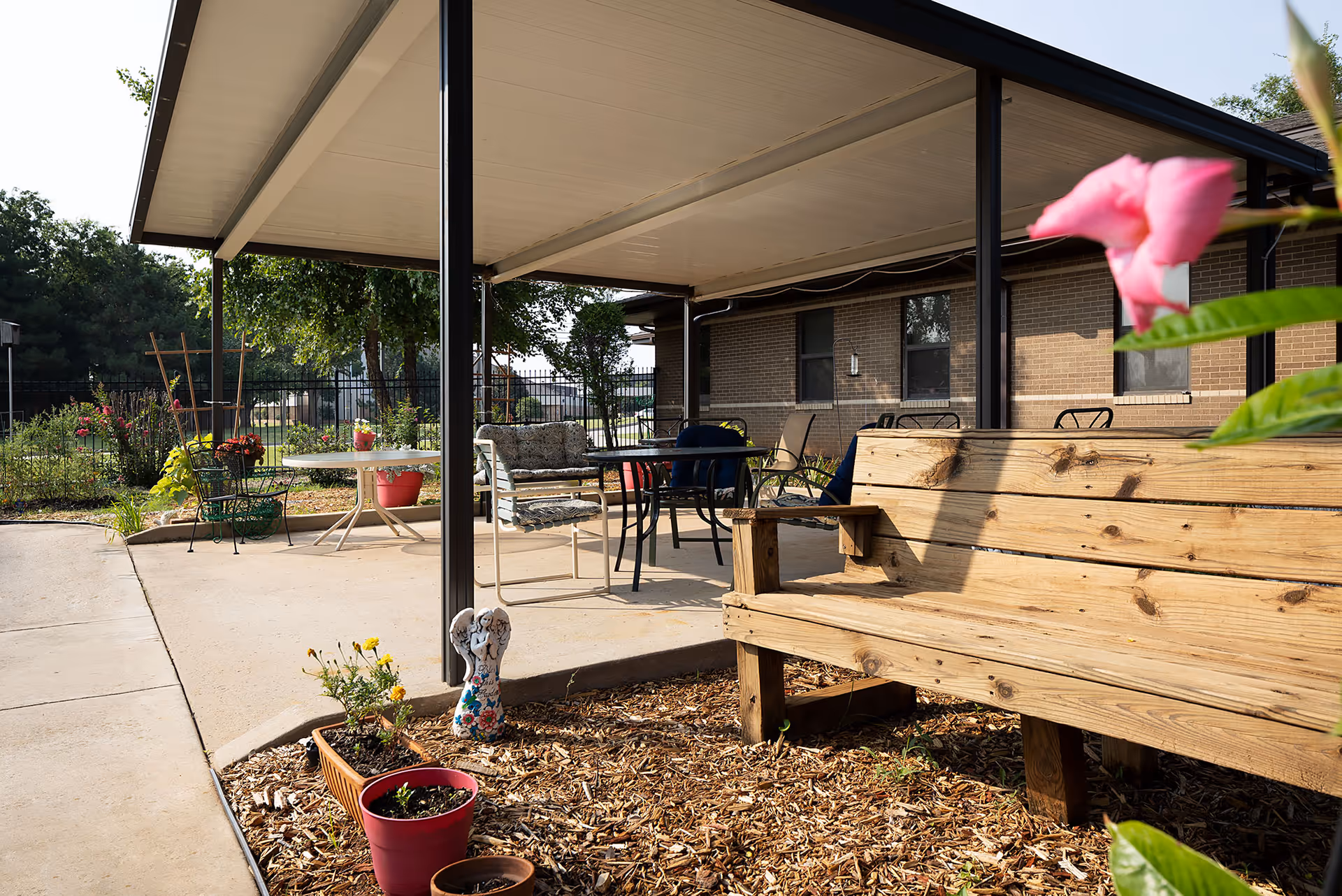 Covered outdoor patio area with a wooden bench, potted plants, and tables and chairs beside a brick building.