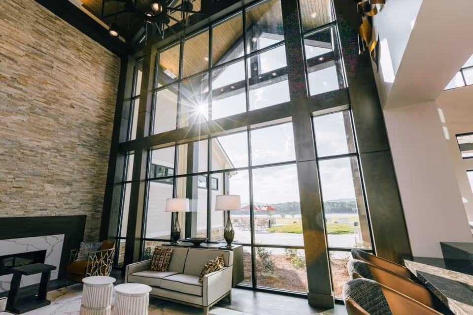 A bright and modern living room area with large floor-to-ceiling windows allowing natural light to flood the space. The room features a beige sofa with patterned cushions, two white round stools, two table lamps on a console behind the sofa, a stone accent wall with a fireplace, and a view of an outdoor patio with red umbrellas and greenery.
