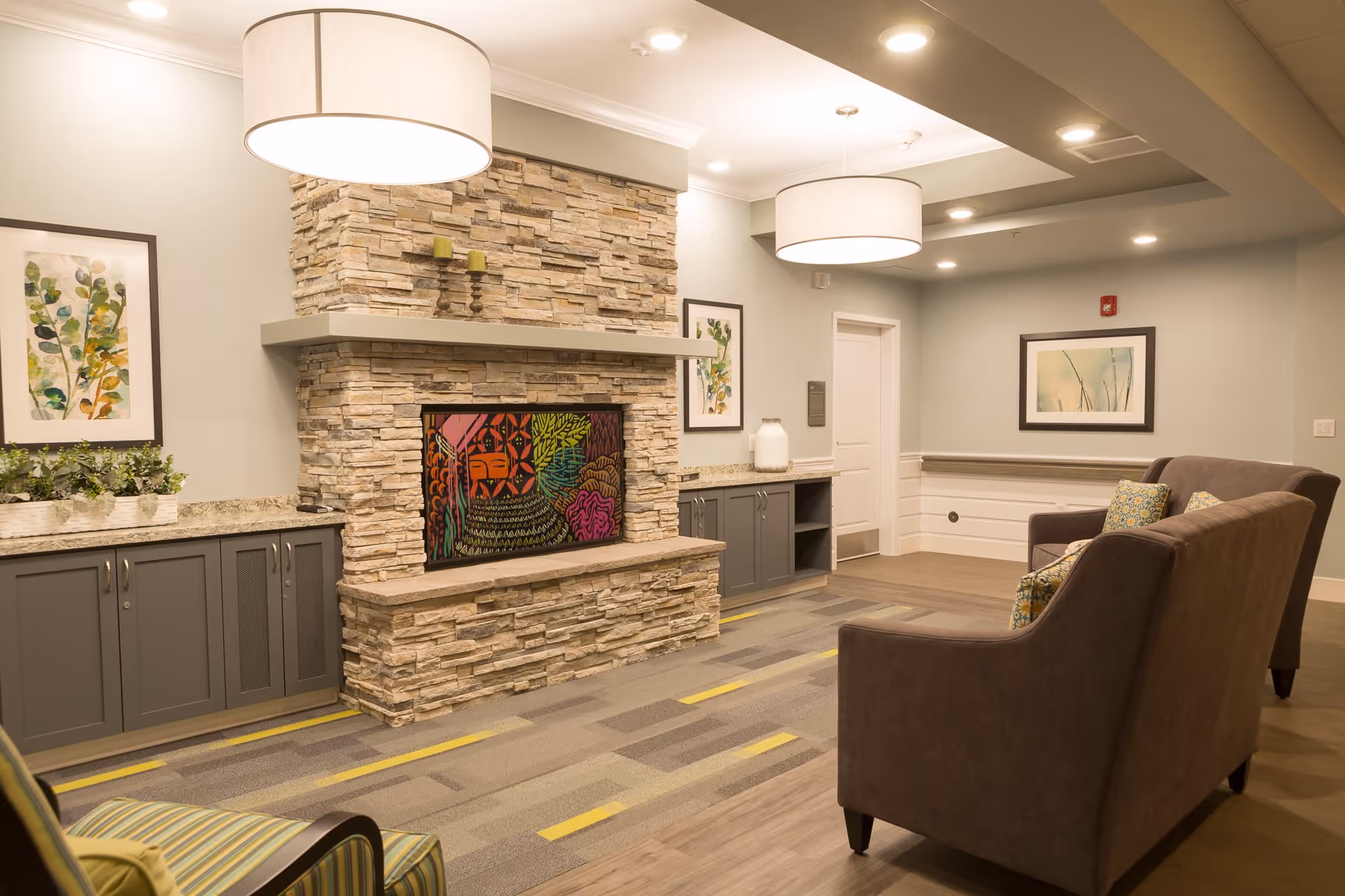 A cozy living room area with a stone fireplace featuring colorful artwork inside the fireplace opening. The room has two brown upholstered sofas with patterned cushions, a striped armchair, and built-in cabinets with granite countertops on either side of the fireplace. The walls are decorated with framed botanical prints, and two large round ceiling lights illuminate the space.