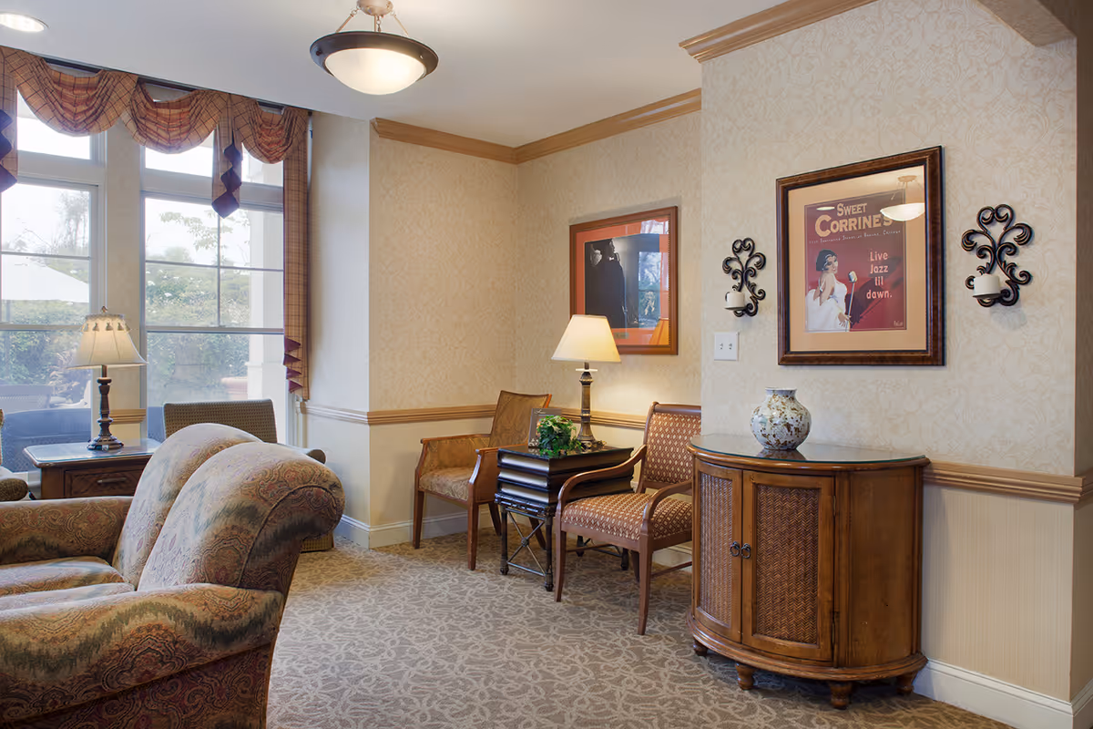 A cozy living room area in a senior living facility with patterned upholstered armchairs, a wooden side table with a lamp, two wooden chairs with a small table between them, a decorative cabinet with a vase on top, framed artwork on the walls, and large windows with plaid valances letting in natural light.