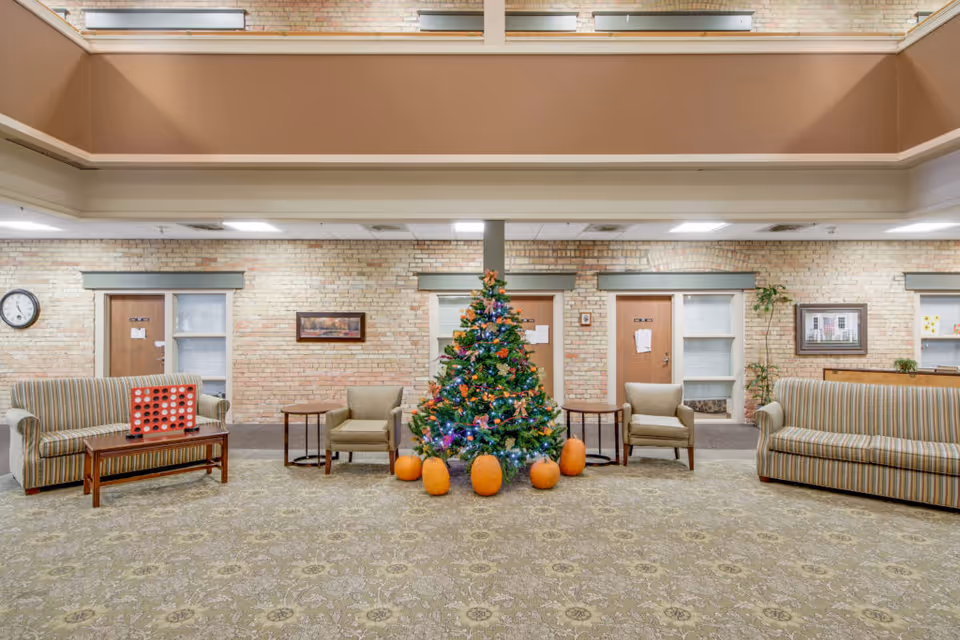 A spacious common area in a senior living facility with a decorated Christmas tree surrounded by pumpkins in the center. The room has two striped sofas, two beige armchairs, wooden side tables, and a coffee table with a Connect Four game on it. The walls are light brick with framed pictures and doors leading to other rooms.