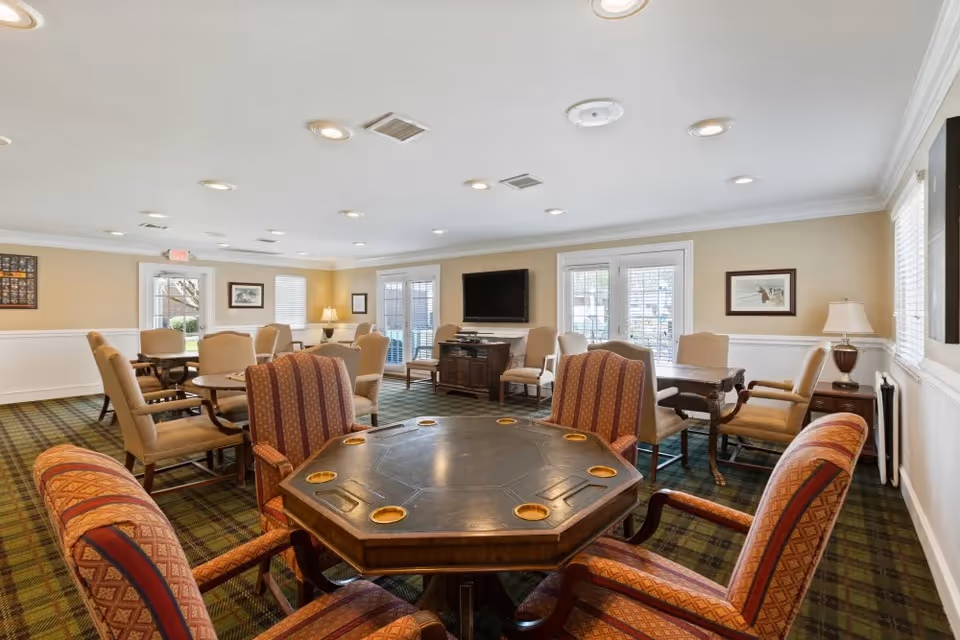 A spacious common room with multiple seating areas including a central octagonal table with eight cushioned chairs, several other tables with beige upholstered chairs, a flat-screen TV mounted on the wall, framed artwork, and large windows letting in natural light.