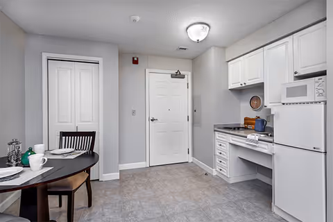 Studio kitchenette and dining area with white cabinets, microwave and refrigerator beside a round table and entry door.
