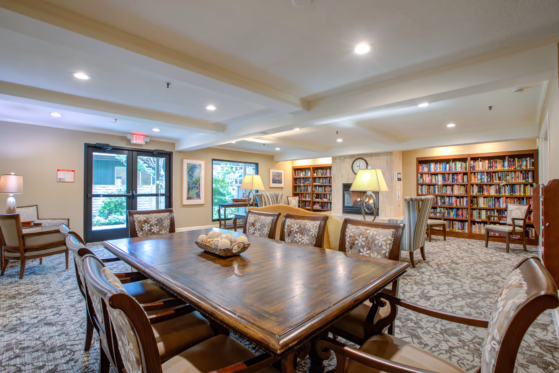 A well-lit common area in a senior living facility featuring a large wooden table with cushioned chairs around it. The room has carpeted floors, a fireplace with a clock above it, bookshelves filled with books, and several lamps providing warm lighting. There are windows and a glass door leading outside, along with additional seating areas with armchairs.