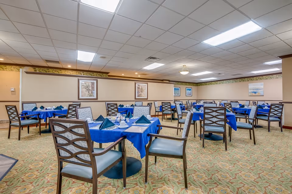 A spacious dining room with multiple tables covered in blue tablecloths, each set with napkins, cups, and utensils. The room has patterned carpet flooring, beige walls with framed artwork, and a white tiled ceiling with fluorescent lighting.