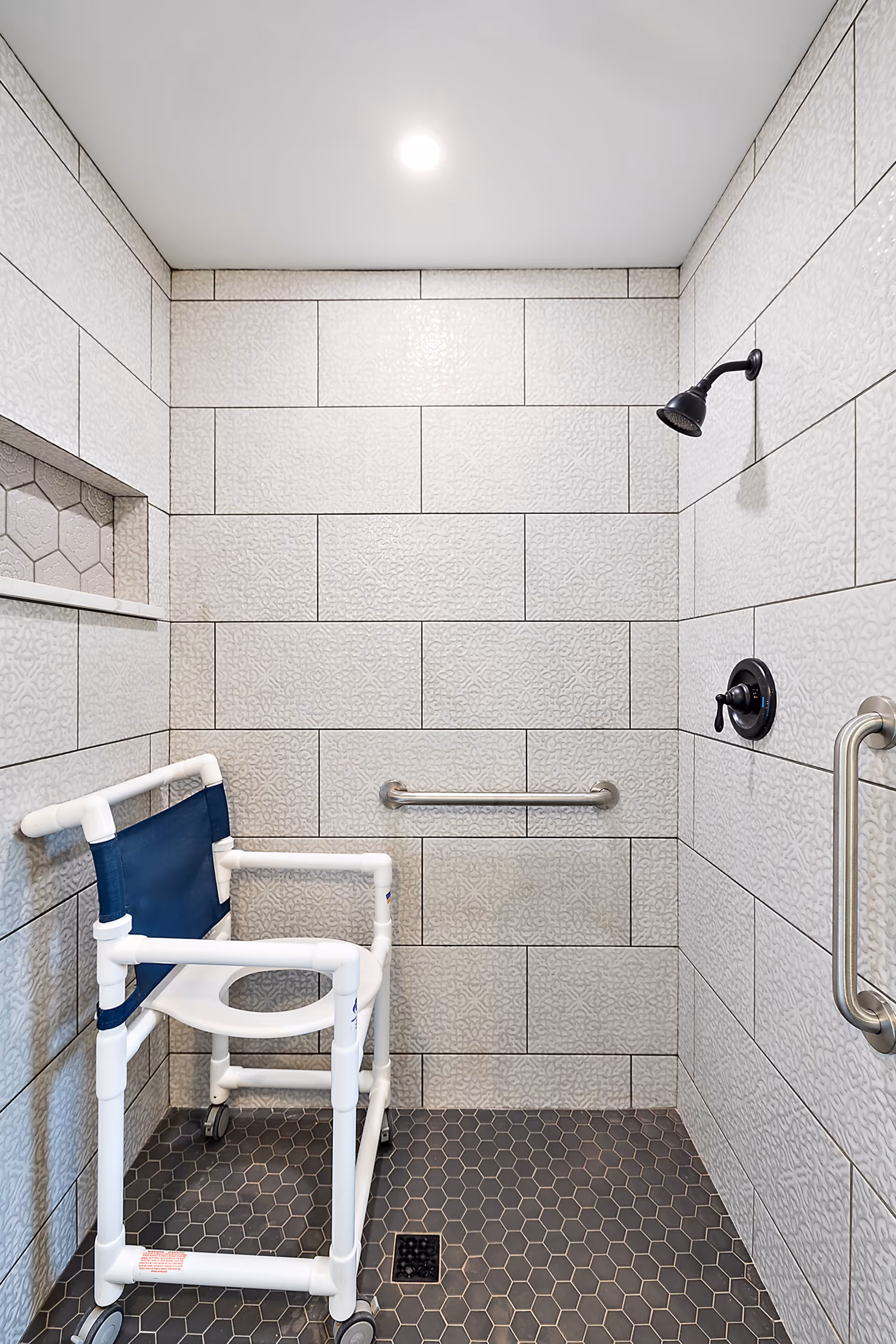 Accessible tiled walk-in shower with a rolling commode/shower chair, grab bars, and an overhead showerhead.