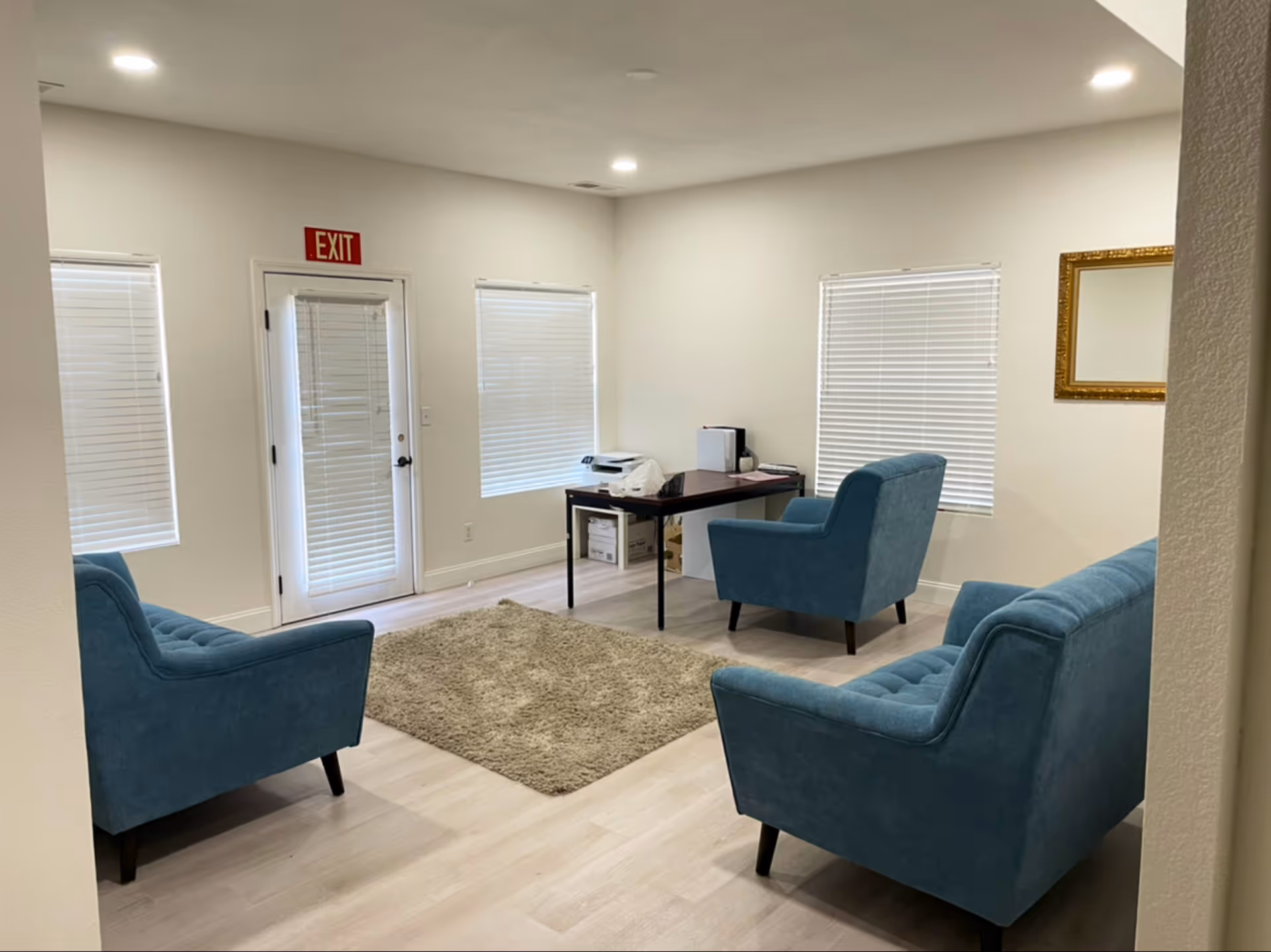 A small office or sitting area with three blue upholstered chairs arranged around a beige rug on a light wood floor. There is a dark wooden desk with office supplies and a printer against the wall, which has three windows with white blinds. A gold-framed mirror hangs on the right wall, and an exit door with a red exit sign is visible on the left side.