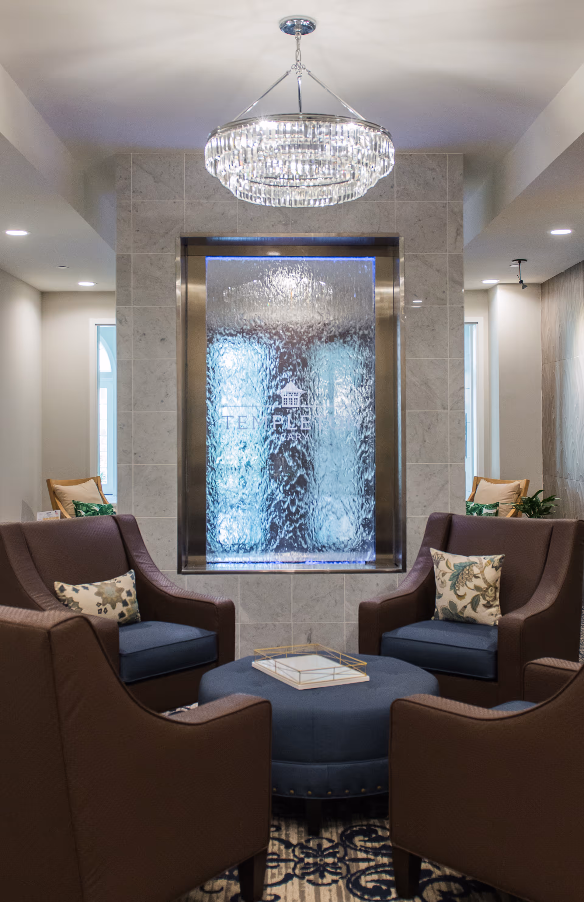 A cozy seating area with four armchairs around a round ottoman in front of a decorative indoor water wall and chandelier.