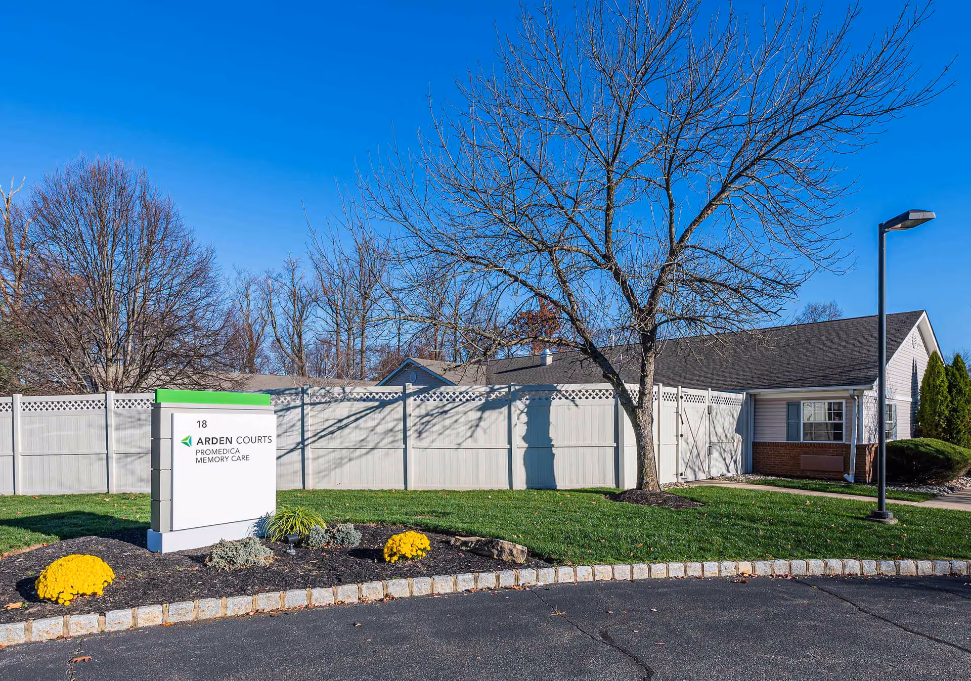 Exterior view of Arden Courts - ProMedica Memory Care Community in Whippany, showing a white fence, a sign with the facility name, a leafless tree, and a building with a gray roof under a clear blue sky.