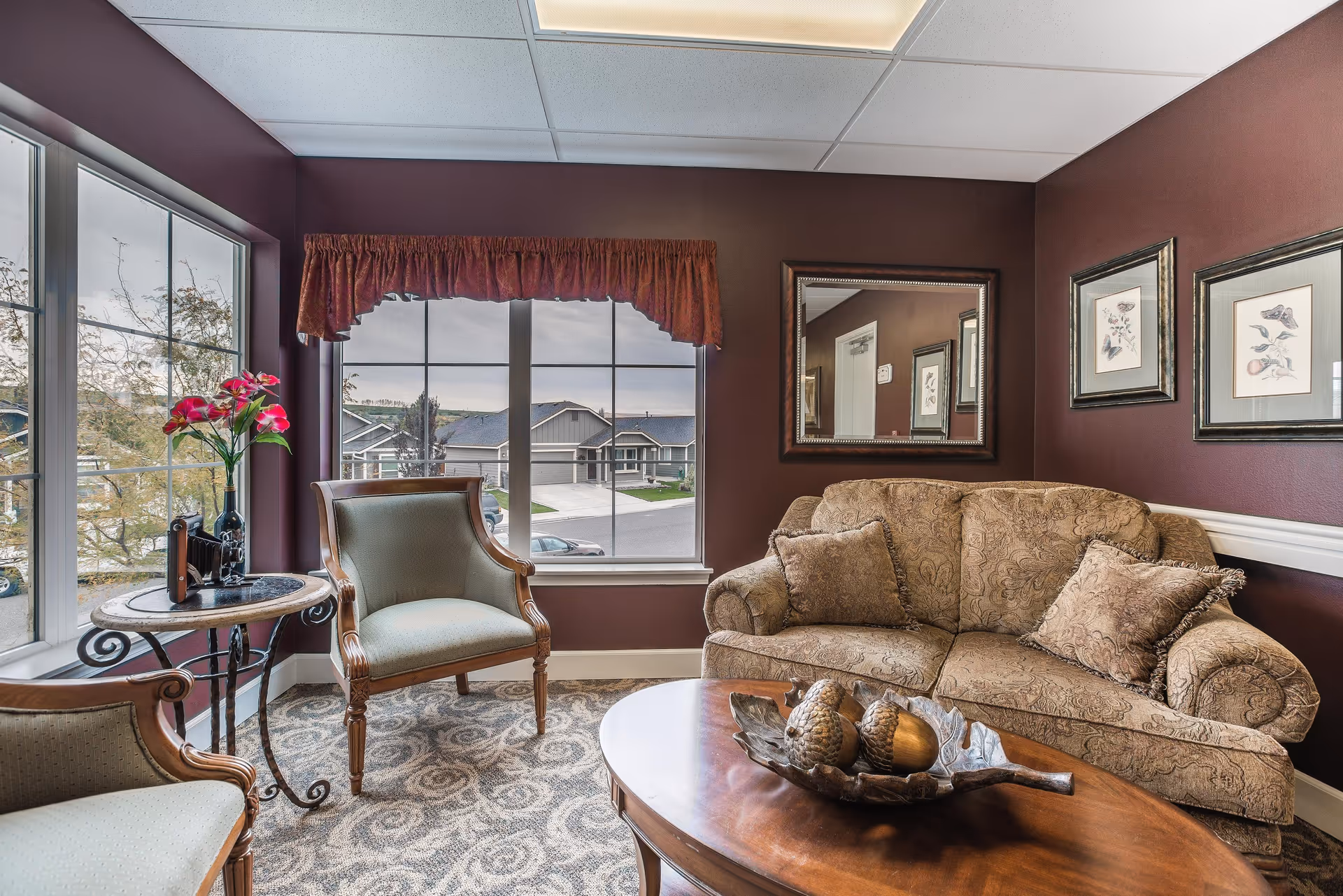 A cozy living room with a patterned beige sofa adorned with matching cushions, two wooden armchairs with light upholstery, a round wooden coffee table with decorative items, and a small round side table with a vase of pink flowers. The walls are painted dark maroon with white trim, decorated with framed botanical prints and a large mirror. Large windows with a maroon valance provide a view of houses and trees outside.