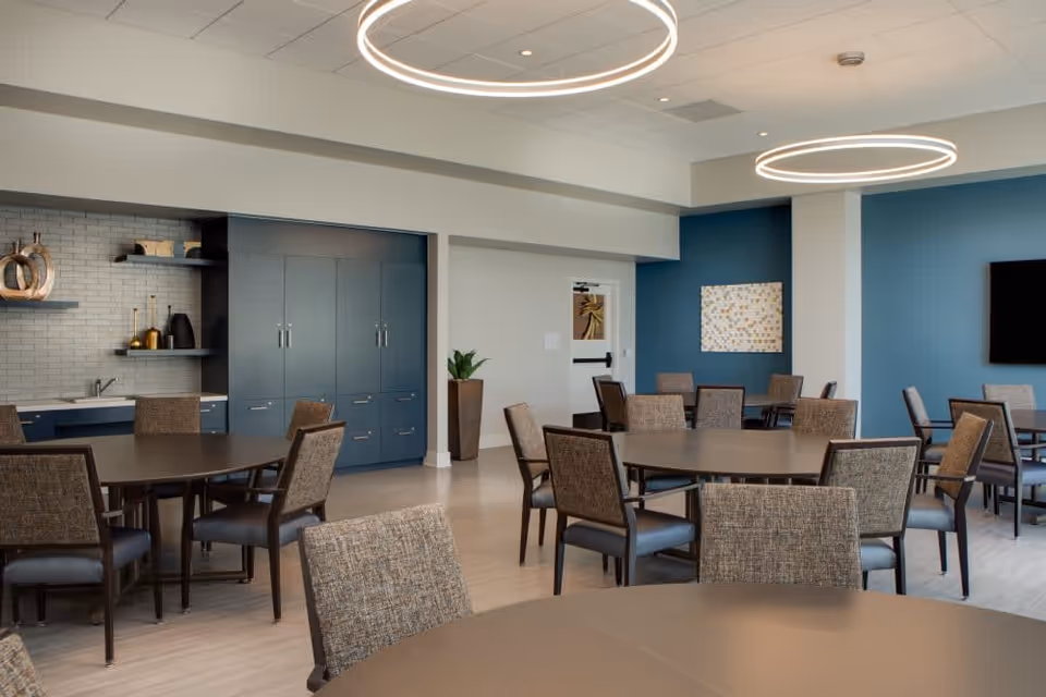 Spacious communal dining room with round tables and upholstered chairs, a kitchenette alcove, blue accent walls and circular ceiling lights.